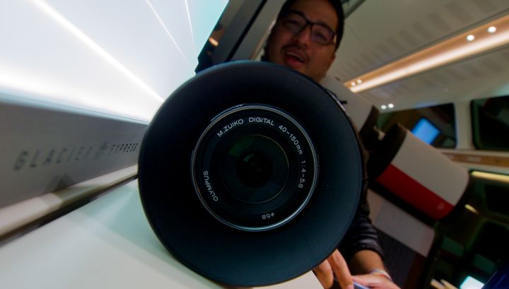 Close-up of a Olympus M.Zuiko lens, on a table in a train, with Jonathan in the background